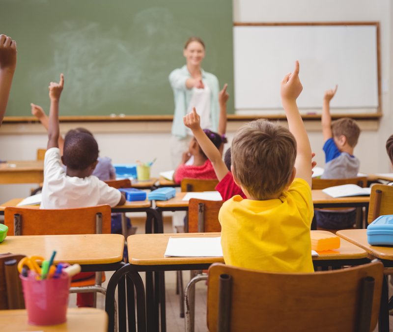 Classroom with children raising their hands. Teacher points to one student.